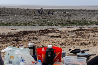 Backs of two people sitting on a beach surrounded by cooking equipment